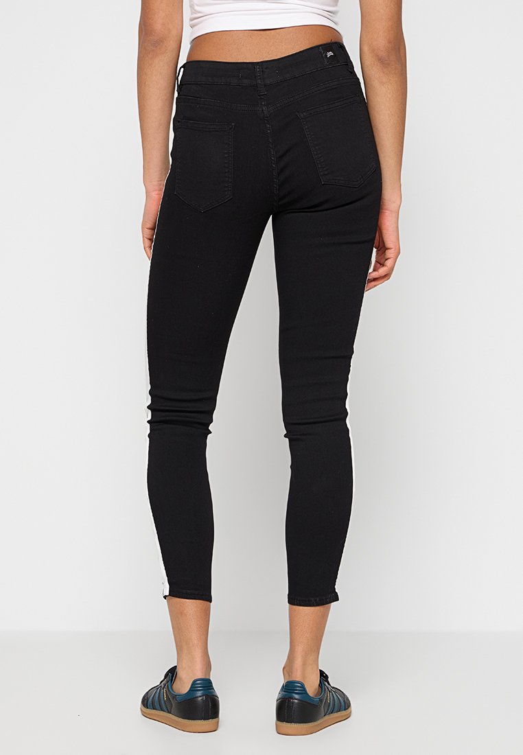 Jean skinny noir en denim extensible, avec une taille haute, deux poches arrière et une silhouette ajustée, associé à des chaussures plates bleues.