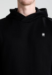 Sudadera negra de algodón, con capucha ajustable y pequeño logo blanco en el pecho. Textura suave y diseño casual.