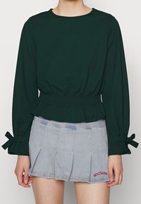 Dunkelgrünes langärmliges Top mit gerafftem Saum und tailliert, kombiniert mit einem hellen, plissierten Denimrock mit roter Stickerei.