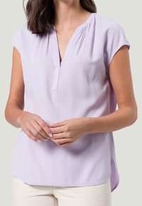 Blouse lilas avec un décolleté en V, des manches courtes et une coupe décontractée. Le tissu est lisse, avec un léger éclat et un ourlet arrondi.