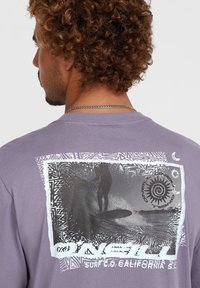 Paarse katoenen t-shirt met een grafische print van een surfer op een bord, omringd door abstracte texturen en patronen. De achterzijde toont een ronde halsontwerp.