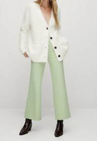 Cardigan bianco e soffice con bottoni neri, tasche frontali, indossato sopra pantaloni a gamba larga verde chiaro e stivaletti marroni. Design minimalista.