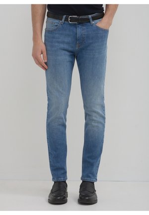 Mężczyzna noszący jasnoniebieskie obcisłe dżinsy, czarny pasek, czarne mokasyny, z jedną ręką w kieszeni, stojący na jednolitym tle.