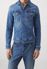 Blå denimjacka med en åtsittande design, knappstängning, två bröstfickor och kontrasterande sömmar. Bärs tillsammans med matchande denimjeans.