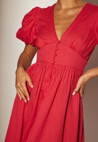 Röd klänning med djup V-ringning, fluffiga korta ärmar, texturerat tyg och knappdetaljer längs fronten. Har en rynkad midja.