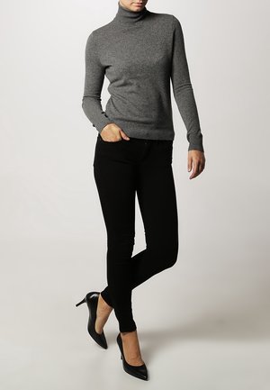 Persoon die een aangesloten grijze coltrui, zwarte skinny broek en zwarte hoge hakken draagt, staat met één hand in de zak.