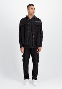 Giacca di velluto a coste nera con tasche frontali, indossata sopra una camicia nera e pantaloni cargo neri. Sneakers bianche completano l'outfit.