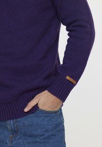 Pullover en maille violet avec des poignets et un ourlet côtelés. Présente une petite étiquette en cuir marron sur le poignet. Associé à un jean en denim bleu.