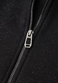 Czarny materiał z subtelnym wzorem w kropki oraz srebrny metalowy suwak. Suwak ma prostokątny uchwyt z logo marki.