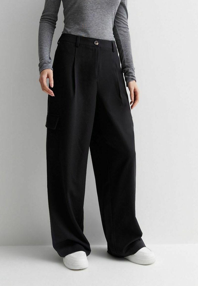 Pantalon large noir en tissu lisse, avec des plis, une fermeture à boutons et une poche cargo latérale. Associé à des baskets blanches.