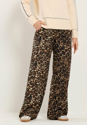 Person iført løstsiddende leopardbukser, beige sweater med sorte syningslinjer og lyse beige sneakers, stående mod en ensfarvet baggrund.