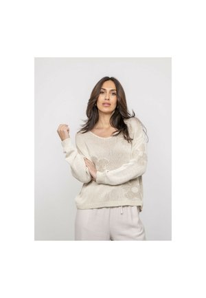 Femme aux longs cheveux foncés portant un pull en tricot beige et un pantalon de couleur claire, posant avec un bras levé devant un fond uni.