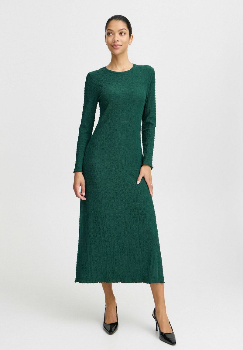 Robe verte à manches longues en tissu texturé avec une coupe droite, atteignant le milieu du mollet, présentant un col rond et des bords côtelés.