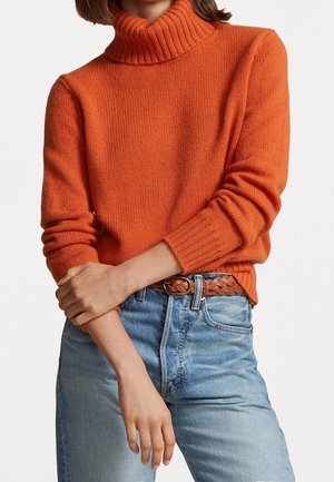 Person trägt einen leuchtend orangenen Rollkragenpullover, der in hellblaue Jeans mit einem geflochtenen braunen Gürtel gesteckt ist, Arme vor der Taille verschränkt.