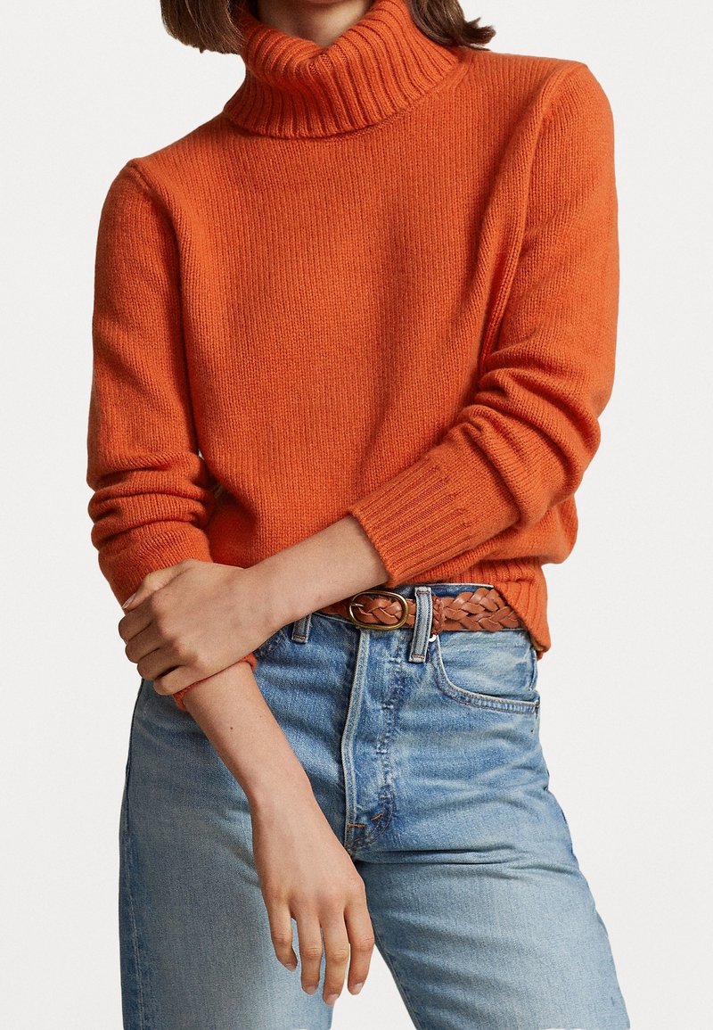 Personne portant un pull à col roulé orange vif rentré dans un jean bleu clair avec une ceinture tressée marron, les bras croisés à la taille.