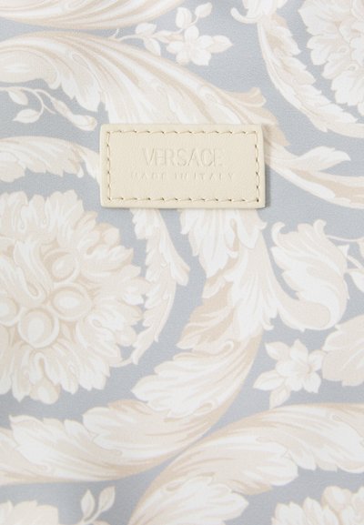 Versace kangas, millel on helehall taust ja keerukad kreemjad lillemustrid, ning õmmeldud beež nahaksilt, millel on reljeefne kiri "VERSACE MADE IN ITALY".