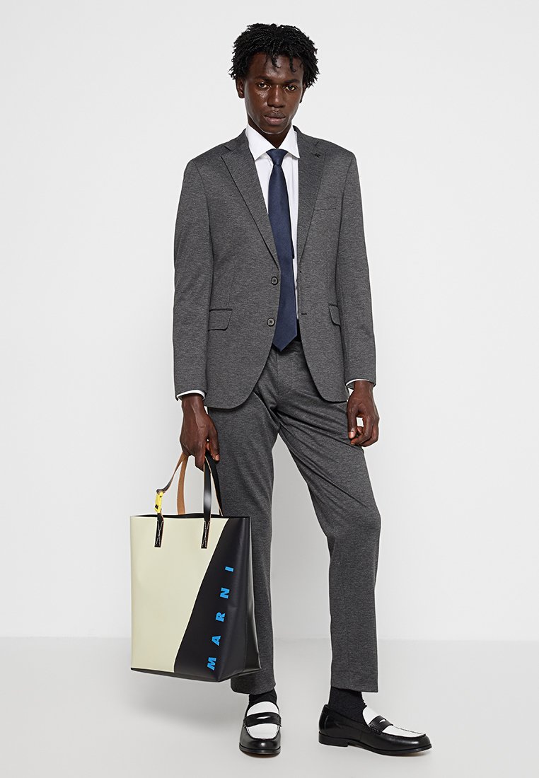 Traje gris a medida con una camisa blanca y una corbata azul marino. Bolsa de mano negra y crema con el texto azul "MARNI". Zapatos en blanco y negro completan el look.