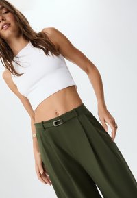 Weißes, verkürztes Tanktop kombiniert mit hochtaillierten, olivgrünen Hosen, die einen Gürtel mit einer silbernen Schnalle und scharfen Falten haben.