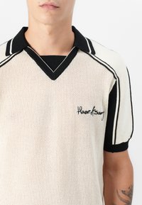 Strickpolo in Creme mit schwarzen Akzenten, mit Mesh-Design, V-Ausschnitt-Kragen und gesticktem Logo auf der Brust.