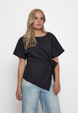 Žena s dlhými blond vlasmi nosí čiernu blúzku s krátkym rukávom a bočným viazaním a svetlomodré rifle s vysokým pásom, stojí proti bielemu pozadiu.