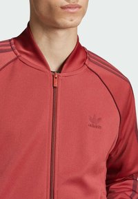 Rote Sportjacke mit hohem Kragen, gerippter Textur, durchgehendem Reißverschluss und drei dunklen Streifen auf jedem Schulter. Mit tonalem Logo.