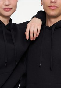 Deux personnes en sweat-shirts noirs à capuche, l'une reposant son bras sur l'épaule de l'autre, toutes deux avec des expressions neutres.