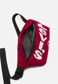 Borsa in tela rossa con logo "Levi's" bianco, apertura con zip, tracolla nera regolabile e fodera interna grigia. Design compatto e strutturato.