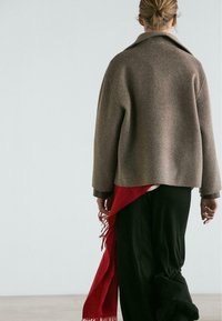 Brązowy wełniany płaszcz z kołnierzem, noszony na czarnych spodniach z czerwonym frędzlowanym akcentem. Tekstura jest miękka; fason jest oversize i casualowy.