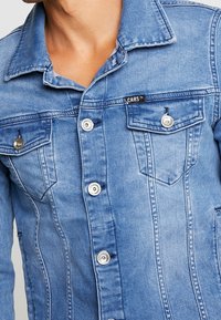 Close-up van een persoon die een blauwe spijkerjack met metalen knopen en borstzakken draagt, met het "CARS" merklabel zichtbaar aan de linkerkant.