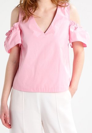 Frau trägt ein pinkfarbenes Cold-Shoulder-Oberteil mit Volantärmeln und weiße, hoch taillierte Hosen, steht mit einer Hand in der Tasche.