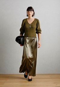 Pull en tricot côtelé vert olive avec des manches bouffantes, associé à une jupe satinée olive et un sac à main noir. Texture lisse et design fluide.