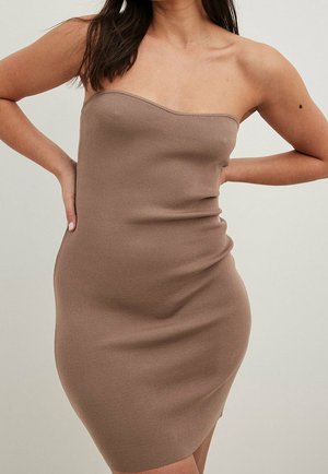 Femme portant une robe beige sans bretelles, ajustée et côtelée, une main sur la hanche et l'autre derrière le dos.