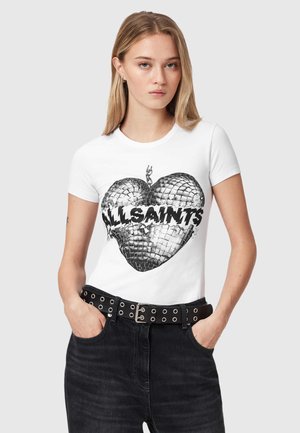 Jeune femme aux longs cheveux blonds portant un t-shirt blanc avec un cœur boule à facettes brisé et le texte "ALLSAINTS", pantalon noir et ceinture.