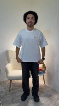 Mann med afro-frisyre iført hvit t-skjorte med grafikk og svarte bukser står i stuen og holder i skjortekanten, med lenestol og sidebord bak seg.