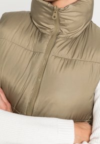 Béžová zateplená vesta s vysokým límcem, zapínáním na zip a hladkou texturou. Bílé pletené rukávy částečně viditelné pod vestou.