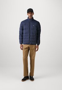 Marineblå pufferjakke med høy krage, med glidelås foran og quiltet tekstur. Kombinert med beige bukser og svarte joggesko.