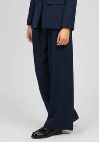 Personne portant un pantalon large marine et une veste assortie avec des mocassins en cuir verni noirs, se tenant contre un fond clair.