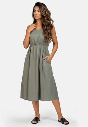 Kobieta o długich falowanych włosach, ubrana w oliwkową, bezrękawnikową sukienkę midi z marszczonym pasem i czarne klapki, pozująca z ręką w kieszeni.