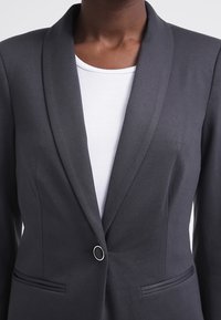 Tmavošedý blejzr s šálovým límcem, zapínáním na jeden knoflík a dvěma předními kapsami; vyrobený z texturované látky, nošený přes bílé tričko.