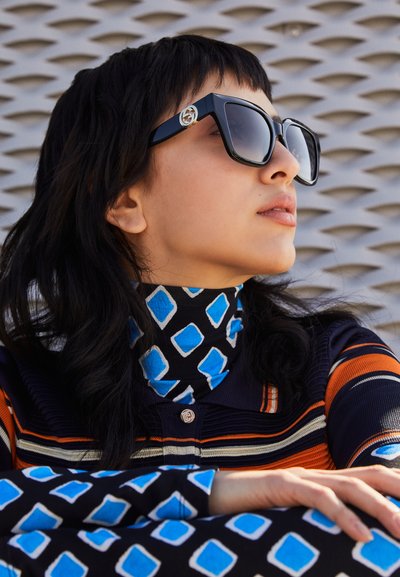 Gafas de sol negras de gran tamaño con un logotipo en el lateral, llevadas con un cuello de tortuga azul y negro que cuenta con formas angulares y rayas.