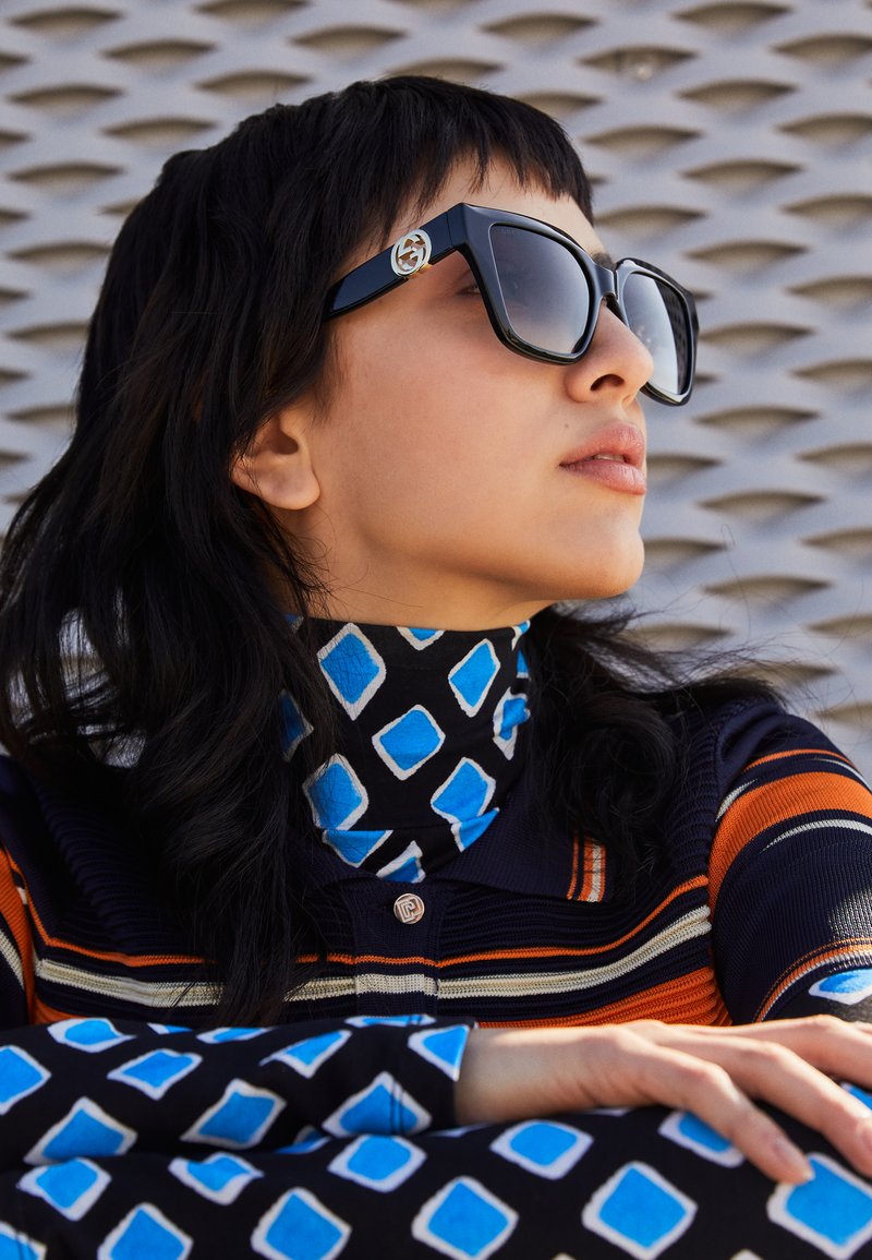 Black oversized sunglasses with a logo on the side, worn with a blue and black patterned turtleneck featuring angular shapes and stripes.