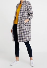 Langer kariertierter Mantel in Blau-, Weiß- und Gelbtönen, mit einem Reverskragen, Seitentaschen und einer klaren Silhouette. Getragen über einem gelben Oberteil.