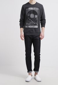 Felpa grigia scura con una grande stampa di un disco in vinile e testo. Abbinata a jeans neri e scarpe slip-on bianche.