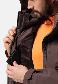 Giacca impermeabile marrone con dettagli in zip e tasche, abbinata a una camicia a maniche lunghe arancione. Presenta polsini regolabili e tessuto testurizzato.
