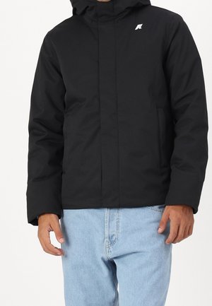 Mann in schwarzer Kapuzenjacke mit Fronttaschen und hellblauen Jeans, steht vor einem schlichten Hintergrund.