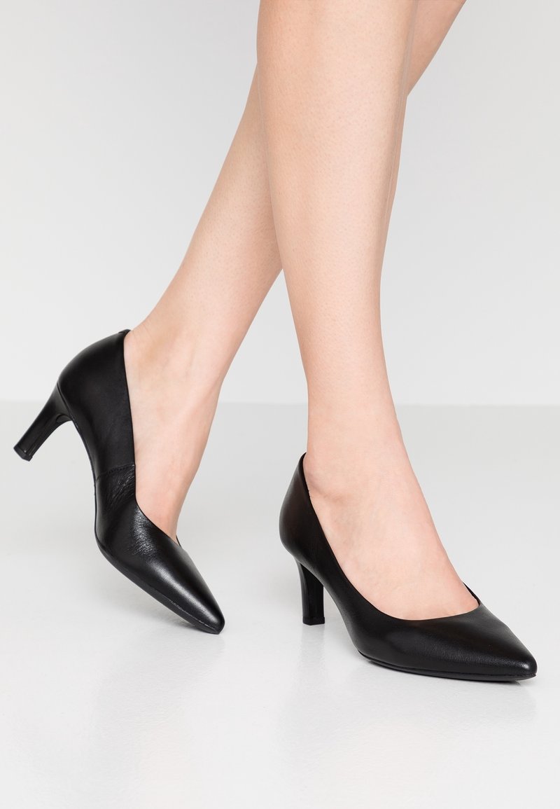 Jambes portant des escarpins en cuir noirs à bout pointu et talons moyens, debout sur un sol gris clair sur un fond uni.