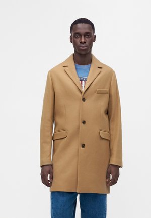 Homme portant un manteau beige à trois boutons sur un t-shirt graphique bleu et un jean bleu, debout devant un fond blanc uni.