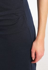 Robe ajustée bleu marine avec une texture lisse, dotée d'une poche latérale et d'un léger froncé près de la taille. Main reposant sur le tissu.