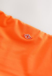 Tissu orange vif avec un petit emblème circulaire argenté présentant un « H » stylisé, fixé près du centre supérieur.