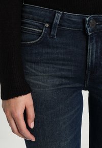 Jeans de denim azul oscuro con corte slim, que cuentan con una cintura de altura media, un desgaste sutil y un diseño estándar de cinco bolsillos. Realzados por un botón dorado.